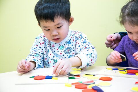 Two children are sitting at a table playing with colorful geometric shapes. One child is wearing a patterned shirt with stars, while the other is dressed in purple. An adult's hand is assisting.